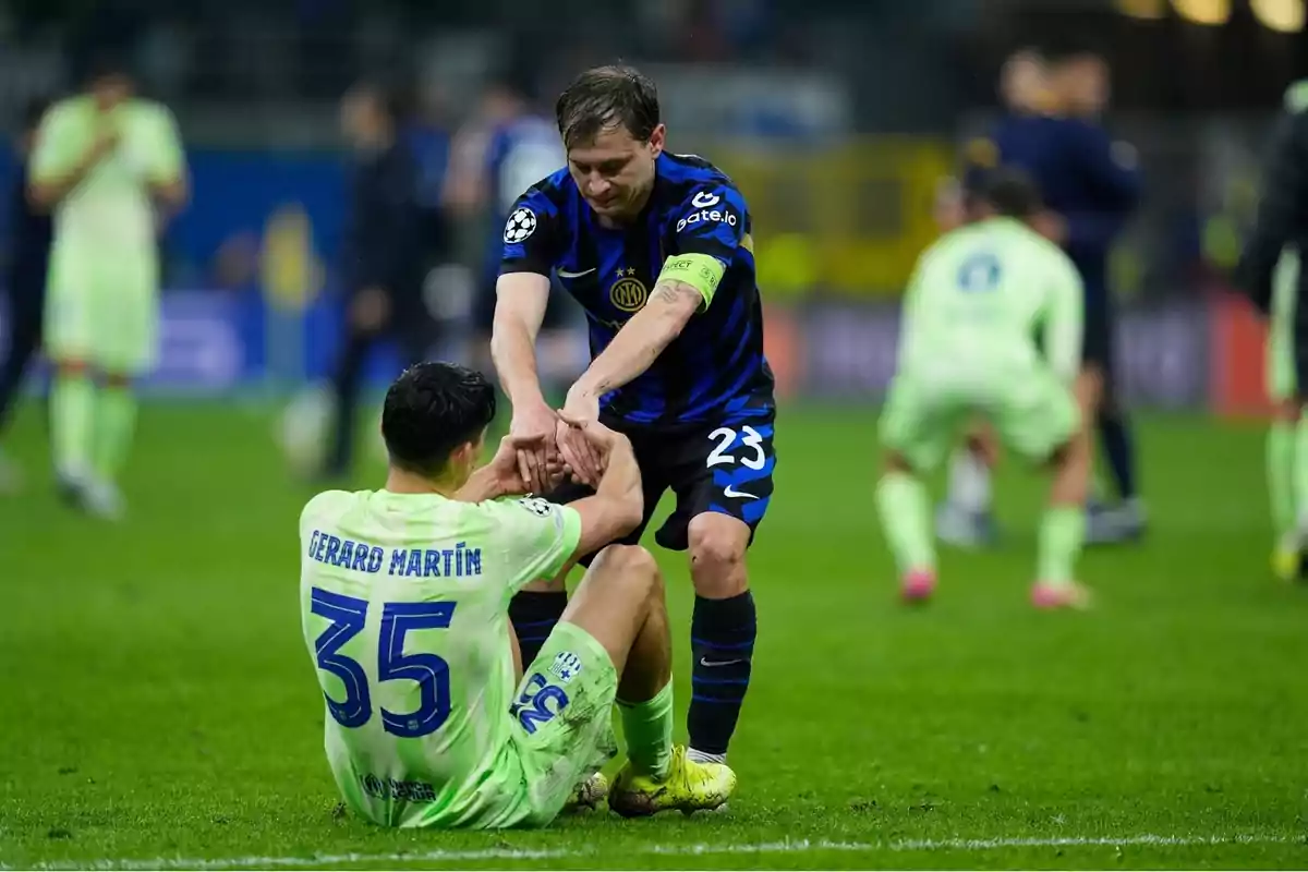 Un jugador de fútbol con uniforme azul ayuda a levantarse a otro jugador con uniforme verde que está sentado en el campo. Un jugador de fútbol con uniforme azul ayuda a levantarse a otro jugador con uniforme verde que está sentado en el campo.