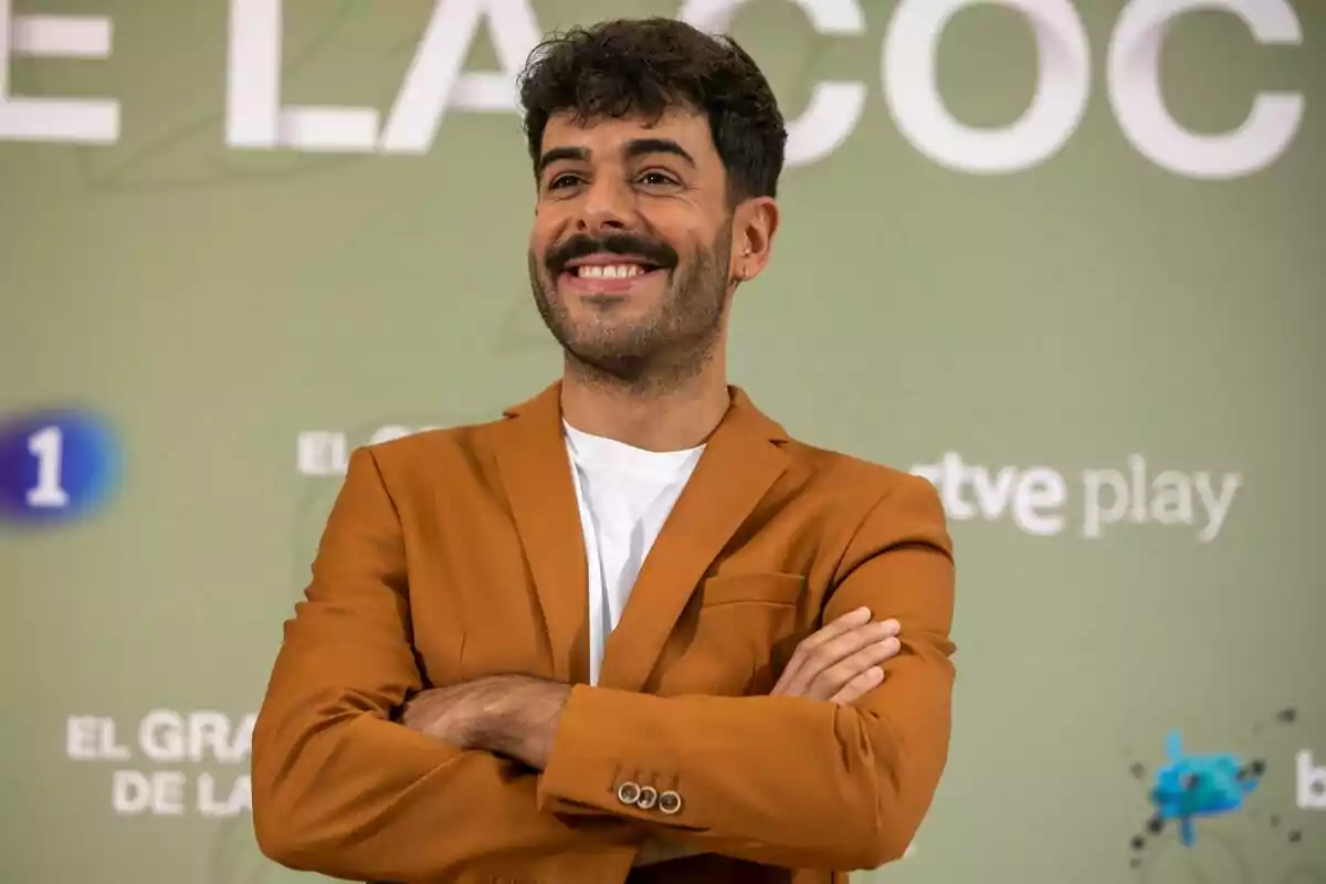 Germán González con chaqueta marrón y camiseta blanca sonriendo con los brazos cruzados frente a un fondo verde con texto en la presentación de El Gran Premio de la Cocina.