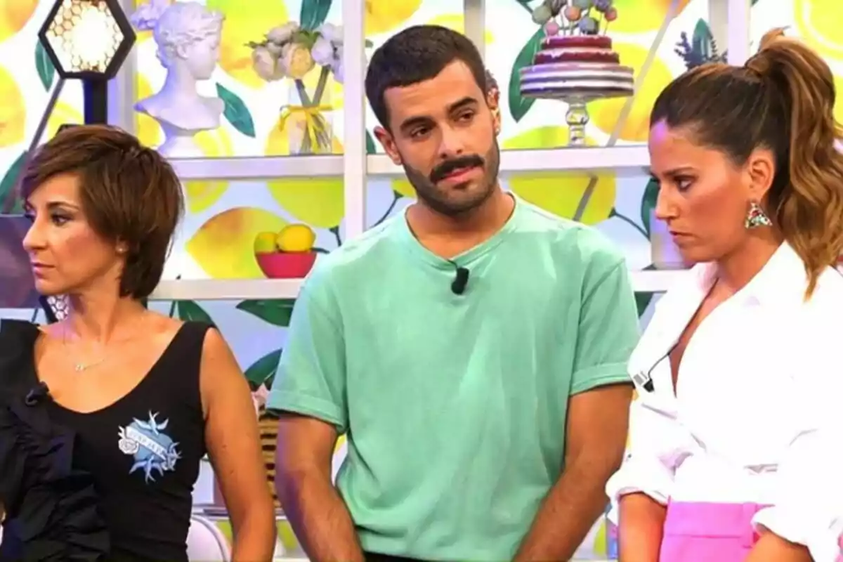 Adela González, Germán González y Nuria Marín están de pie en un set de televisión decorado con motivos de frutas y pasteles, una mujer con cabello corto y oscuro lleva una camiseta negra, un hombre con barba y bigote viste una camiseta verde, y otra mujer con el cabello recogido lleva una blusa blanca y una falda rosa en el plató de Sálvame.