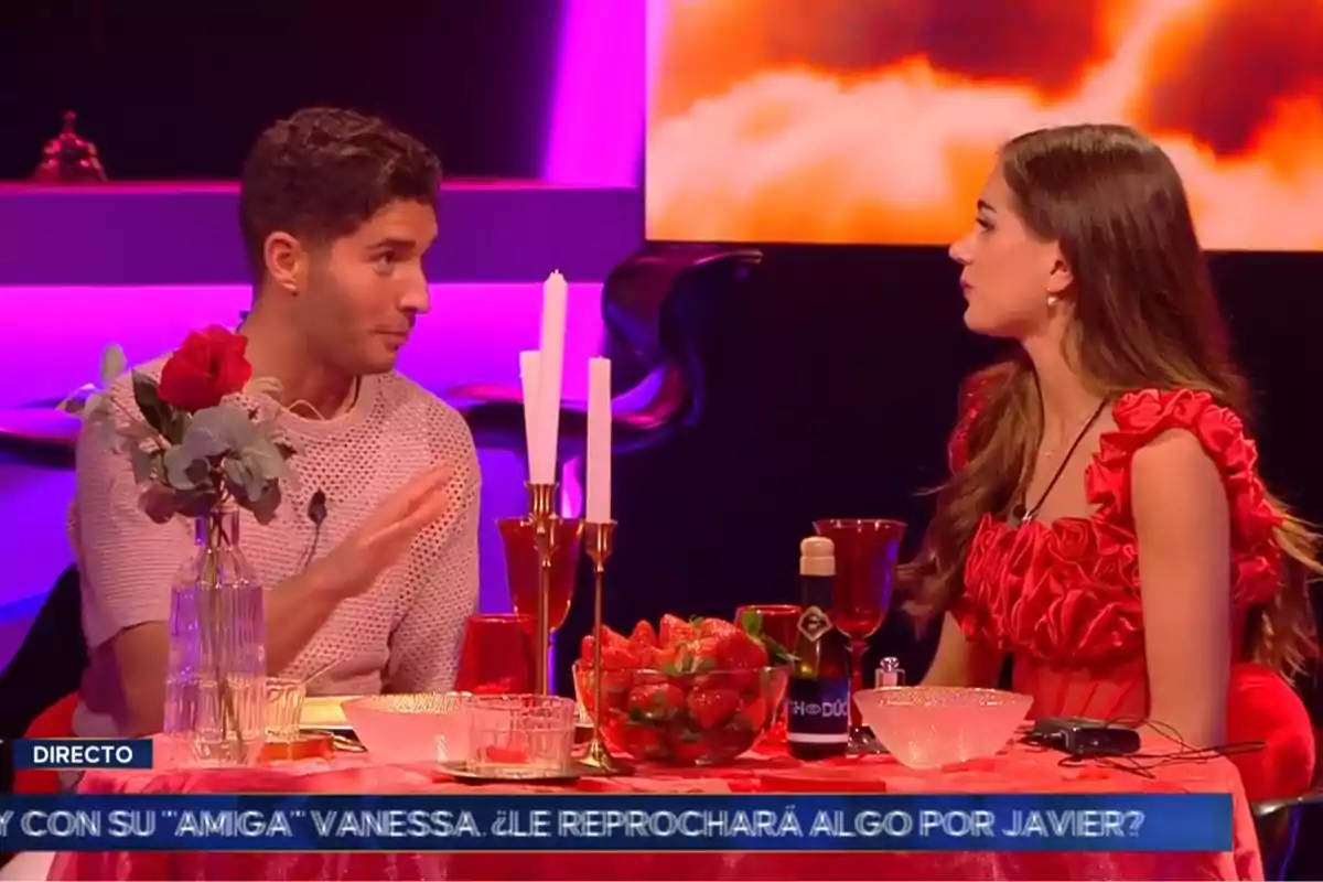 Álex Ghita y Maica conversan en un set de televisión decorado con tonos rojos y morados, con una mesa adornada con velas, una rosa y fresas. Captura de 'GH Dúo' Álex Ghita y Maica conversan en un set de televisión decorado con tonos rojos y morados, con una mesa adornada con velas, una rosa y fresas. Captura de 'GH Dúo'