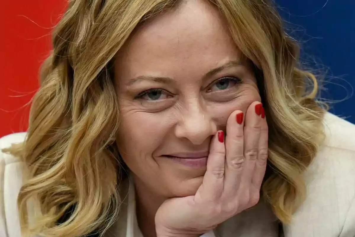 Una persona con cabello rubio rizado y uñas pintadas de rojo sonríe mientras apoya su rostro en su mano.