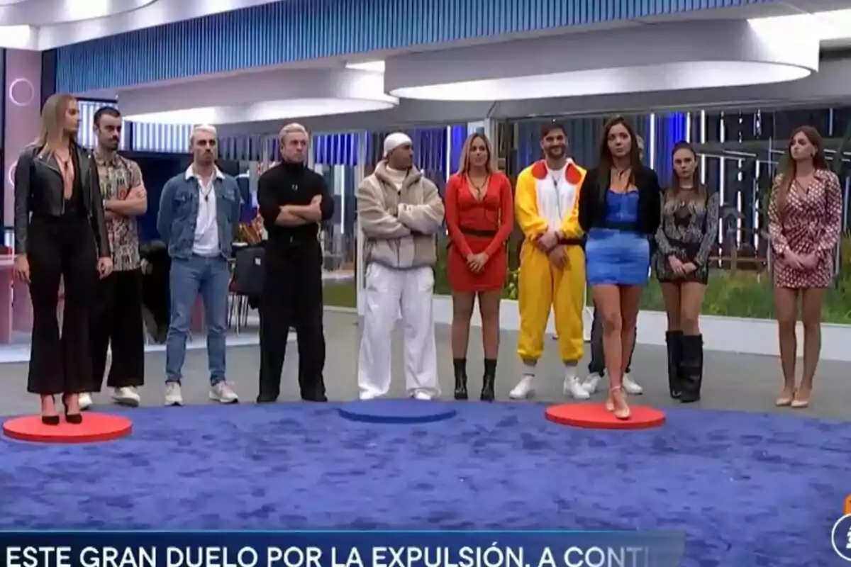 Un grupo de concursantes de Gran Hermano en pie en la casa, algunas sobre círculos de colores en el suelo, con un letrero en la parte inferior que menciona un duelo por la expulsión.