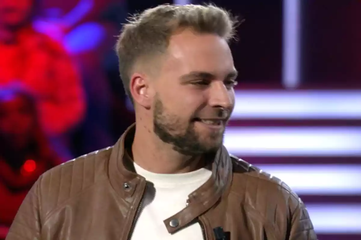 José María Almoguera con chaqueta de cuero marrón y camiseta blanca sonríe mientras mira hacia un lado en un entorno con luces de fondo. Captura de 'Gran Hermano'