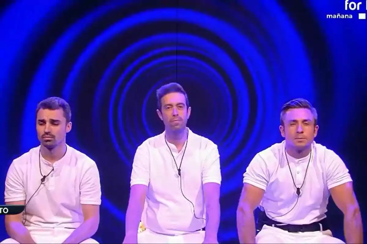 Jorge, Javi y Juan sentados con camisas blancas y micrófonos, frente a un fondo azul con un patrón de círculos concéntricos en la sala de expulsión de Gran Hermano.