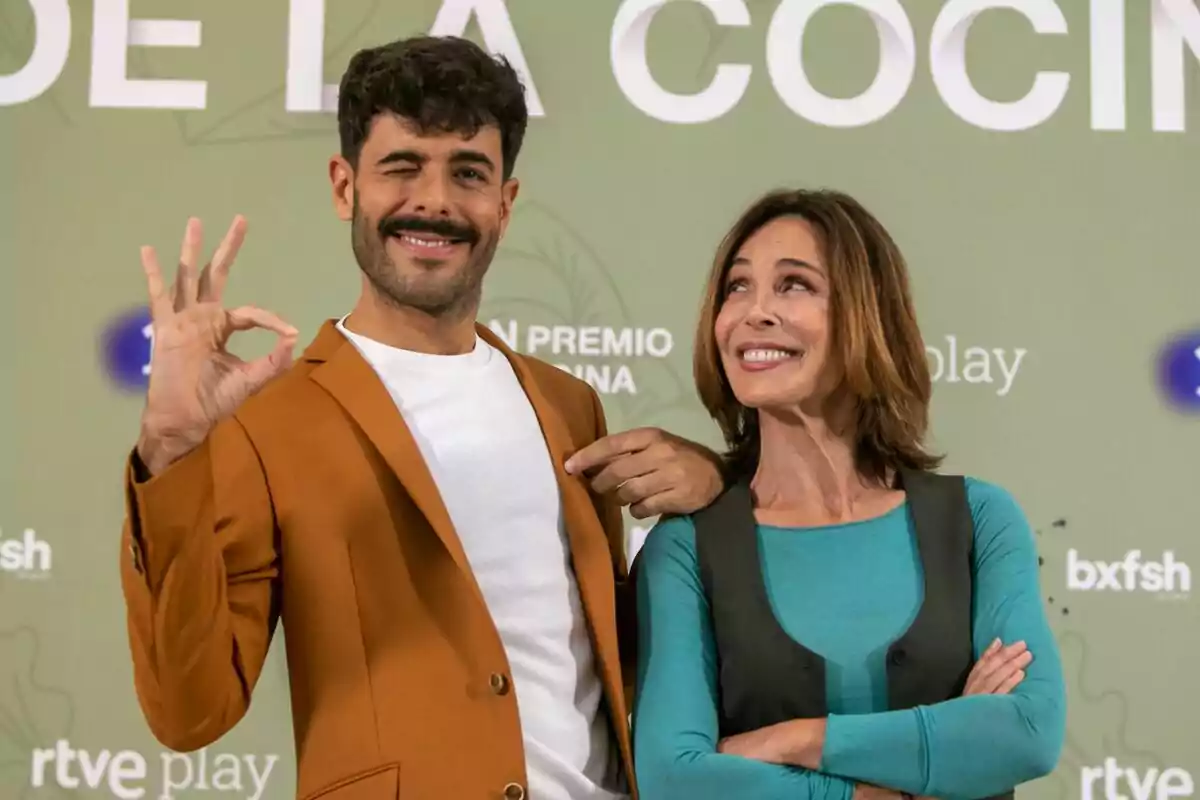 Germán González y Lydia Bosch posan frente a una pared verde con texto, una de ellas hace un gesto de "OK" con la mano y guiña un ojo mientras la otra sonríe en la presentación de El Gran Premio de la Cocina.