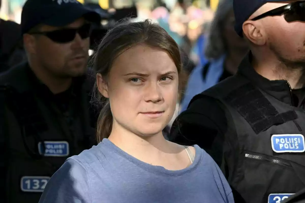 Greta Thunberg con camiseta azul claro rodeada de policías con gafas de sol y chalecos negros en un evento al aire libre. Greta Thunberg con camiseta azul claro rodeada de policías con gafas de sol y chalecos negros en un evento al aire libre.