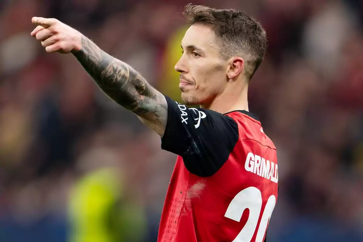 Un jugador de fútbol con tatuajes en el brazo señala hacia adelante durante un partido, vistiendo una camiseta roja con el número 20. Un jugador de fútbol con tatuajes en el brazo señala hacia adelante durante un partido, vistiendo una camiseta roja con el número 20.