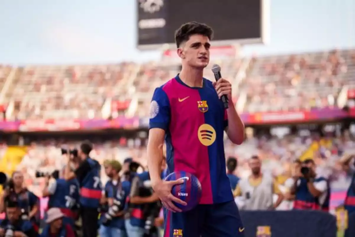 Un jugador de fútbol con el uniforme del FC Barcelona sostiene un micrófono y un balón en un estadio lleno de espectadores y fotógrafos. Un jugador de fútbol con el uniforme del FC Barcelona sostiene un micrófono y un balón en un estadio lleno de espectadores y fotógrafos.
