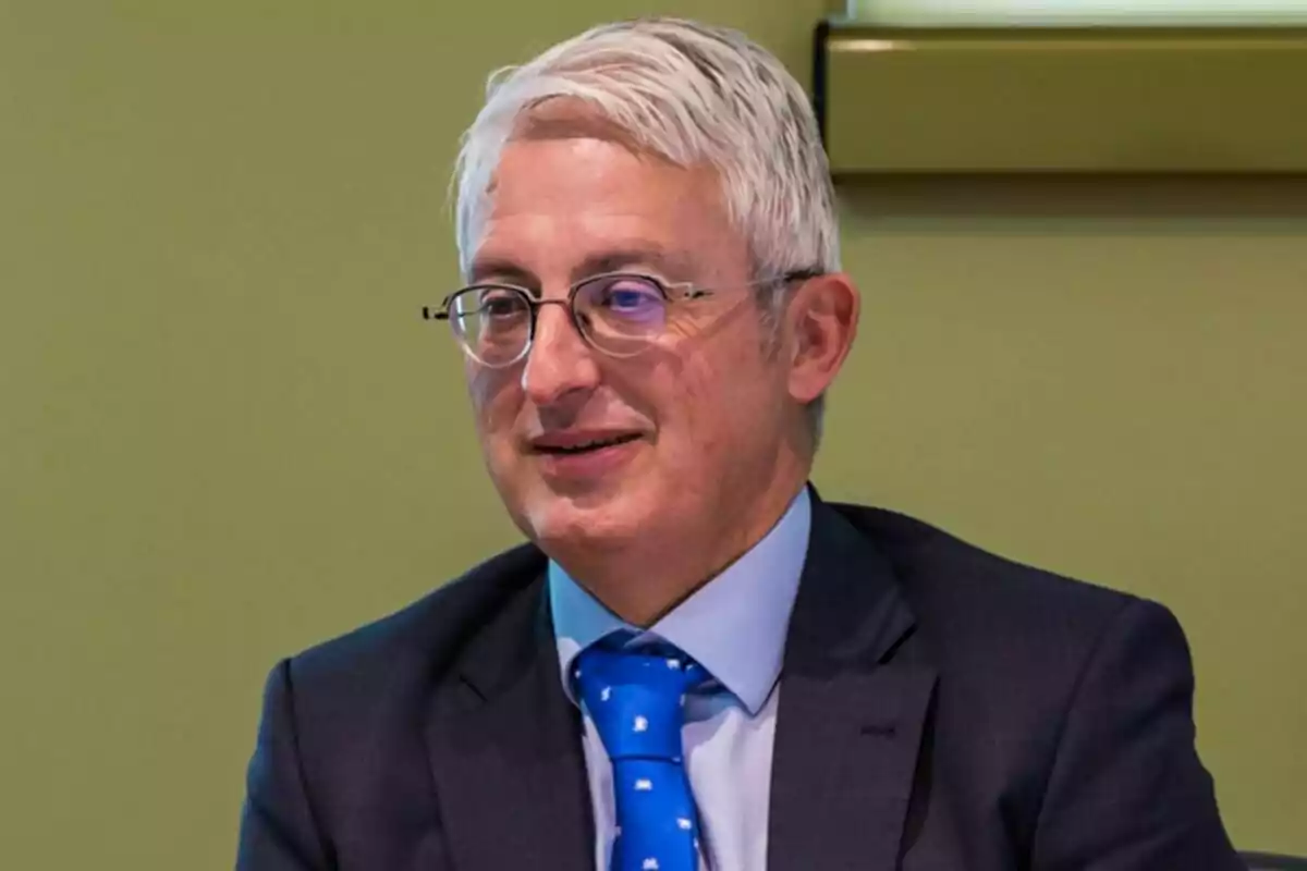 Hombre de cabello canoso y gafas con traje oscuro y corbata azul sentado frente a una pared verde Hombre de cabello canoso y gafas con traje oscuro y corbata azul sentado frente a una pared verde