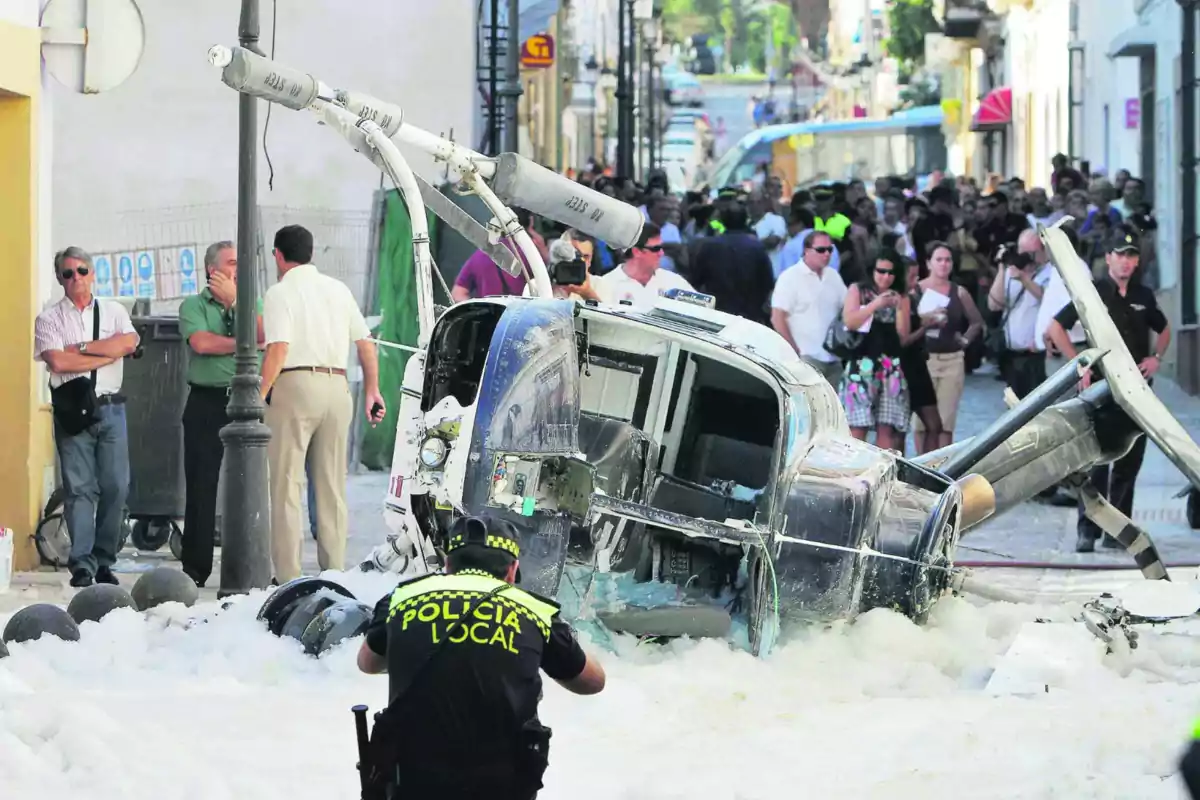 Un helicóptero accidentado yace en una calle rodeado de espuma mientras un policía local observa la escena y varias personas se agrupan alrededor. Un helicóptero accidentado yace en una calle rodeado de espuma mientras un policía local observa la escena y varias personas se agrupan alrededor.