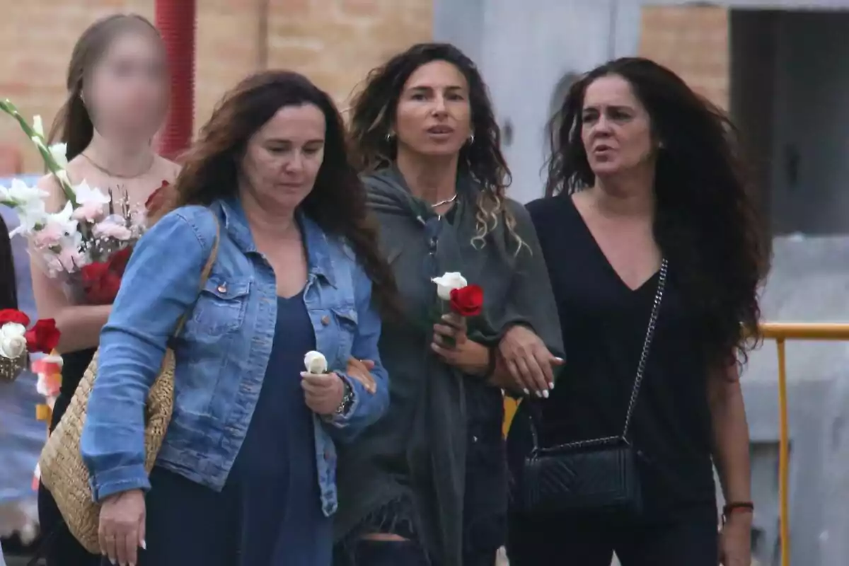 Tres hermanas de Olga Moreno caminan juntas, una de ellas lleva una chaqueta de mezclilla y una bolsa de paja, otra lleva una sudadera gris y una rosa roja, y la tercera lleva una blusa negra y un bolso negro; al fondo, una persona desenfocada sostiene un ramo de flores. Tres hermanas de Olga Moreno caminan juntas, una de ellas lleva una chaqueta de mezclilla y una bolsa de paja, otra lleva una sudadera gris y una rosa roja, y la tercera lleva una blusa negra y un bolso negro; al fondo, una persona desenfocada sostiene un ramo de flores.