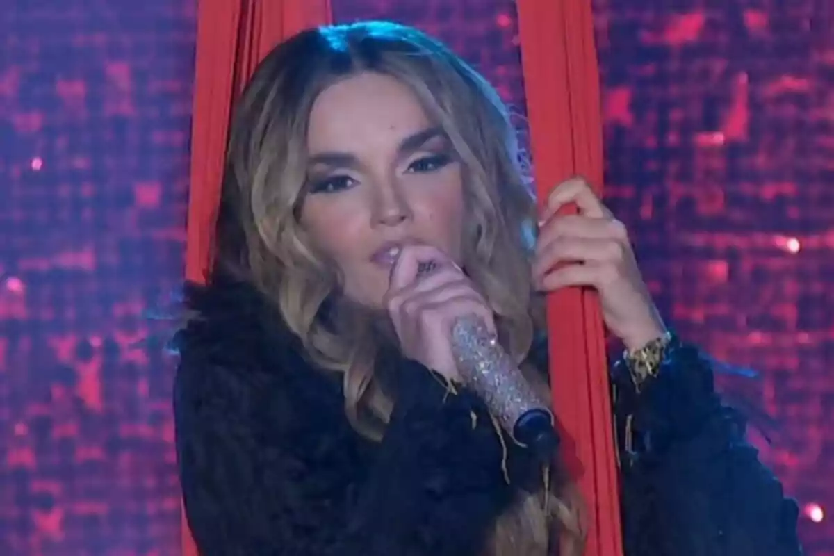 Melody cantando en el escenario de El Hormiguero con micrófono brillante y sosteniéndose de telas rojas Melody cantando en el escenario de El Hormiguero con micrófono brillante y sosteniéndose de telas rojas