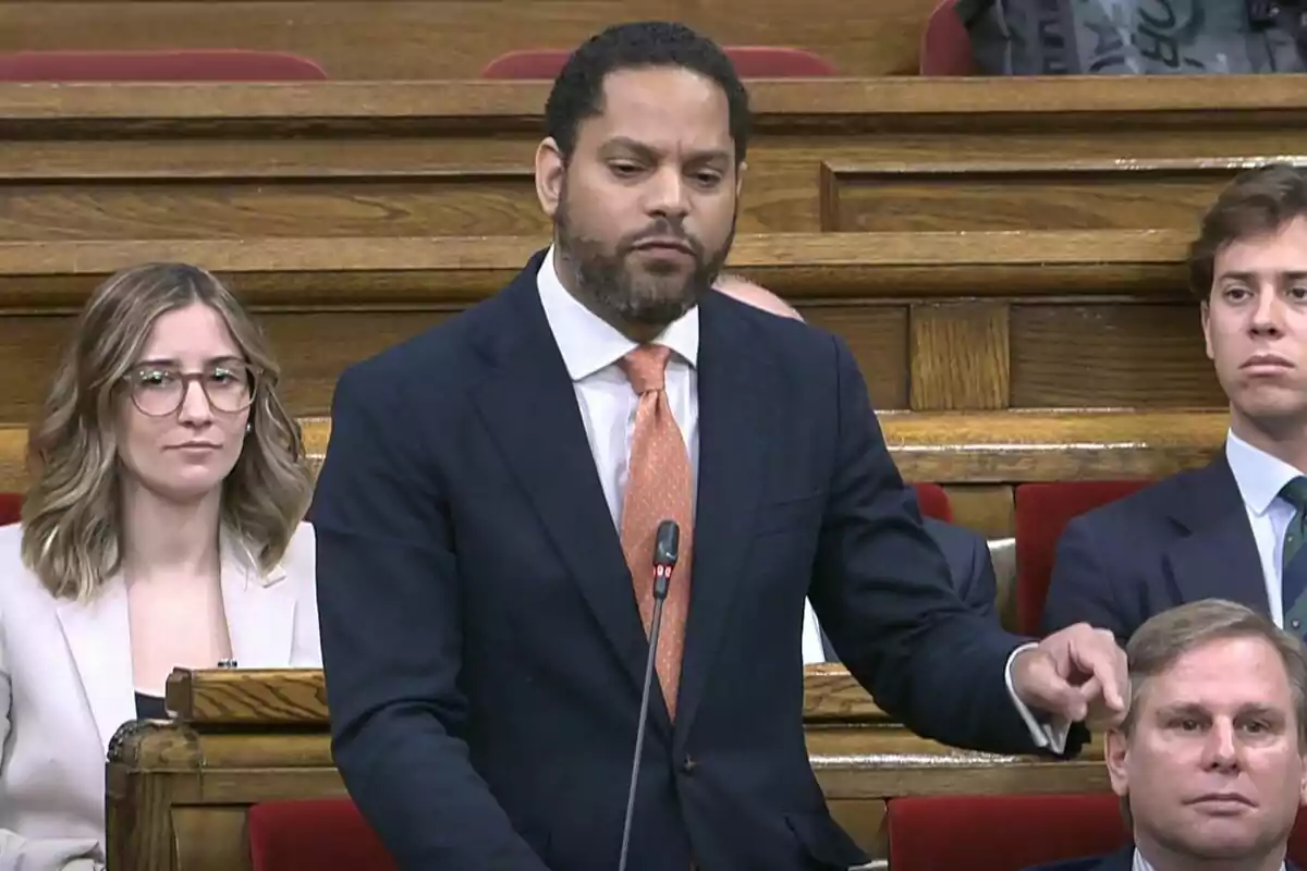Un hombre de pie en un entorno formal, posiblemente un parlamento, hablando mientras otras personas lo observan.