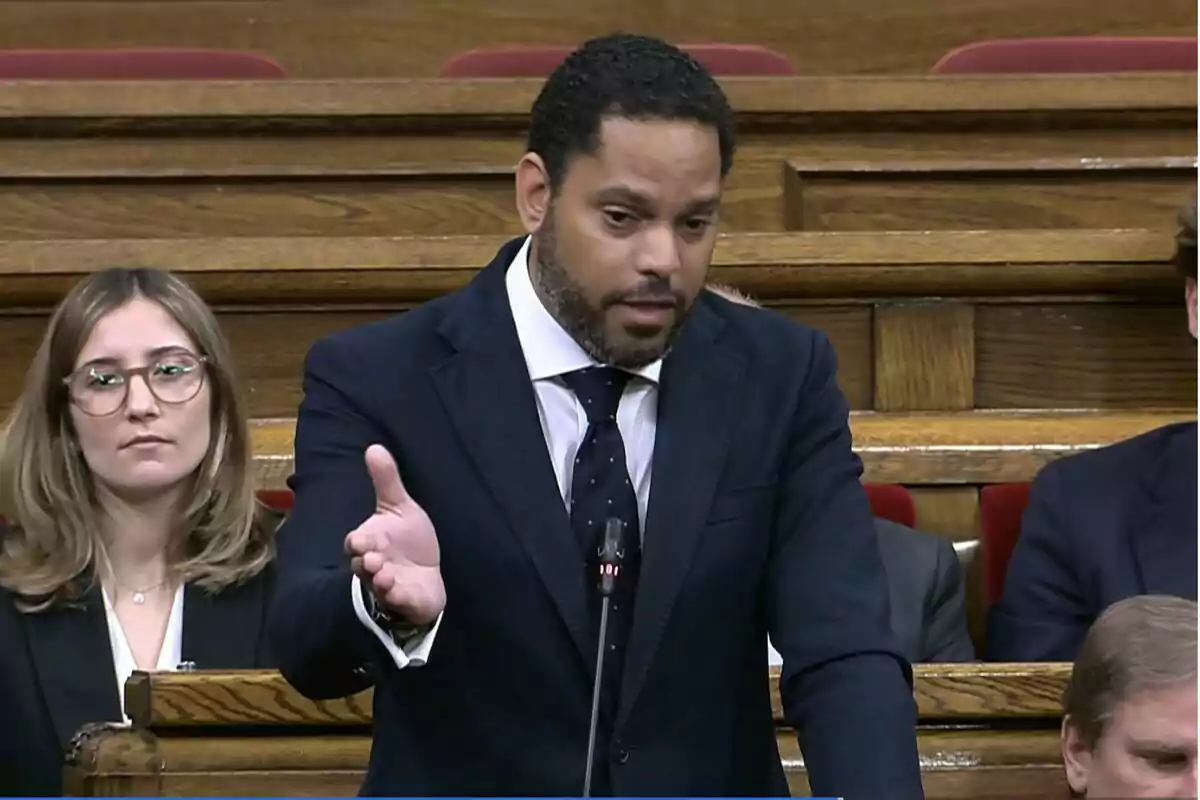 Un hombre de traje oscuro habla gesticulando con la mano mientras está de pie en un recinto parlamentario con otras personas sentadas a su alrededor Un hombre de traje oscuro habla gesticulando con la mano mientras está de pie en un recinto parlamentario con otras personas sentadas a su alrededor