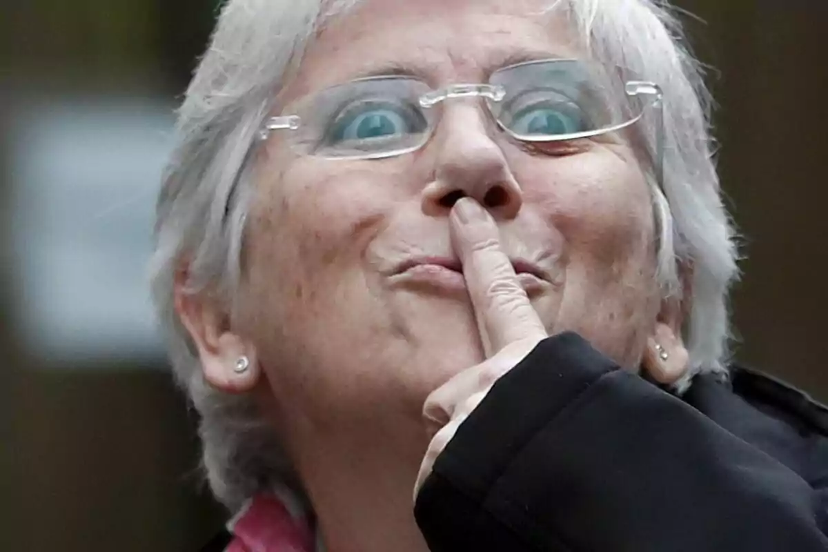 Persona mayor con cabello canoso y gafas, haciendo un gesto de silencio con el dedo índice sobre los labios.