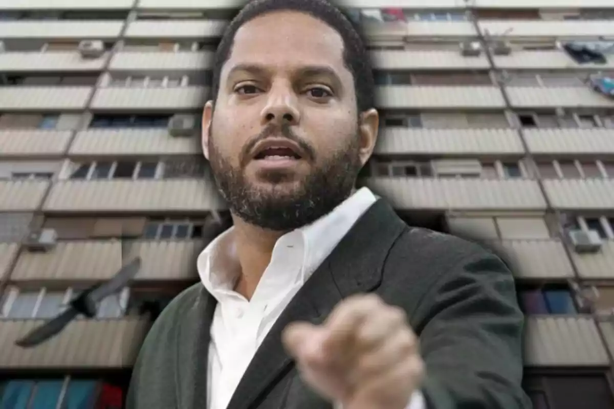 Hombre con barba y traje oscuro señalando hacia la cámara con un edificio de apartamentos de fondo.