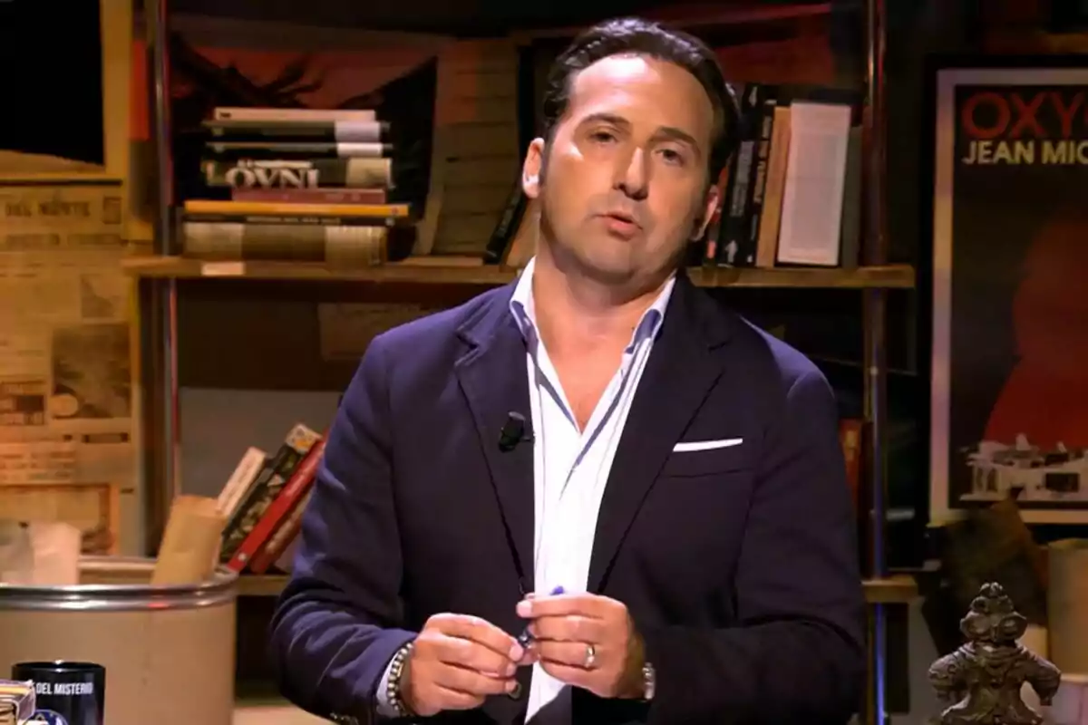 Iker Jiménez de cabello oscuro con saco azul marino y camisa blanca de pie en un estudio con estanterías llenas de libros y carteles en la pared en Cuarto Milenio Iker Jiménez de cabello oscuro con saco azul marino y camisa blanca de pie en un estudio con estanterías llenas de libros y carteles en la pared en Cuarto Milenio