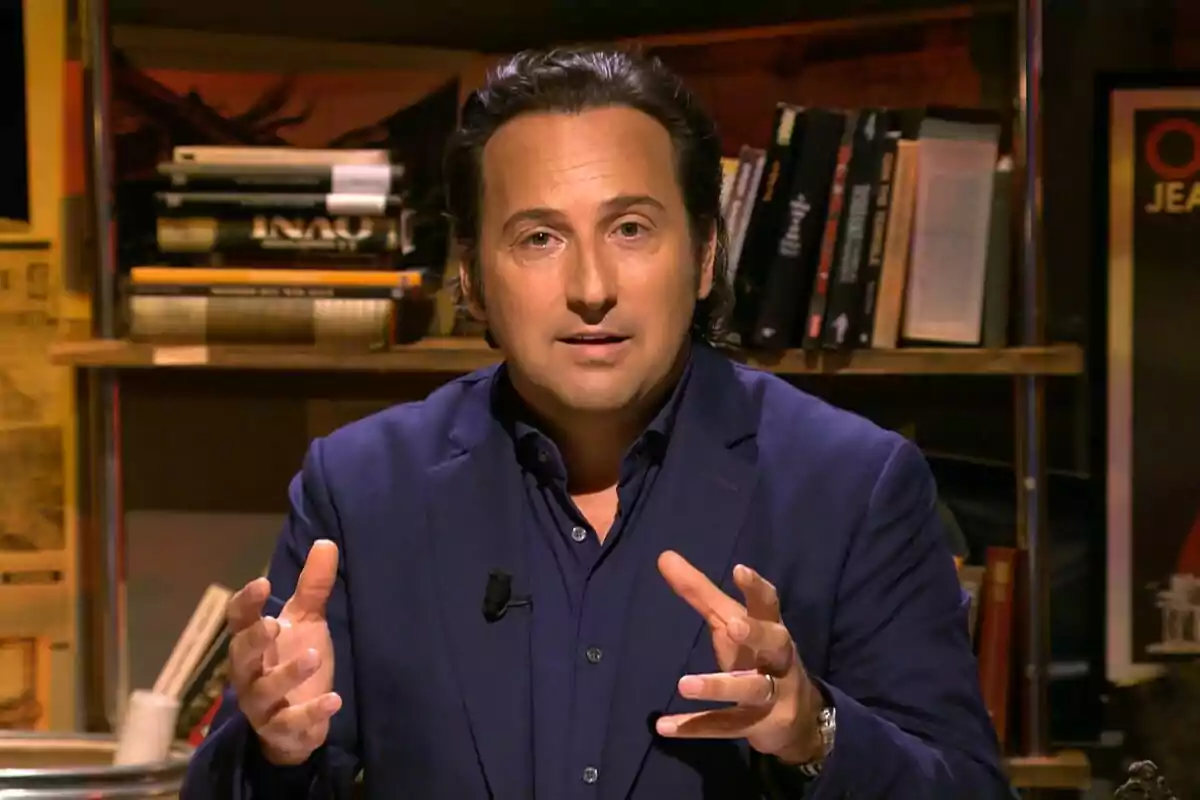 Captura de Iker Jiménez con traje azul oscuro está hablando y gesticulando con las manos, mientras está sentado frente a una estantería llena de libros en el final de Cuarto Milenio