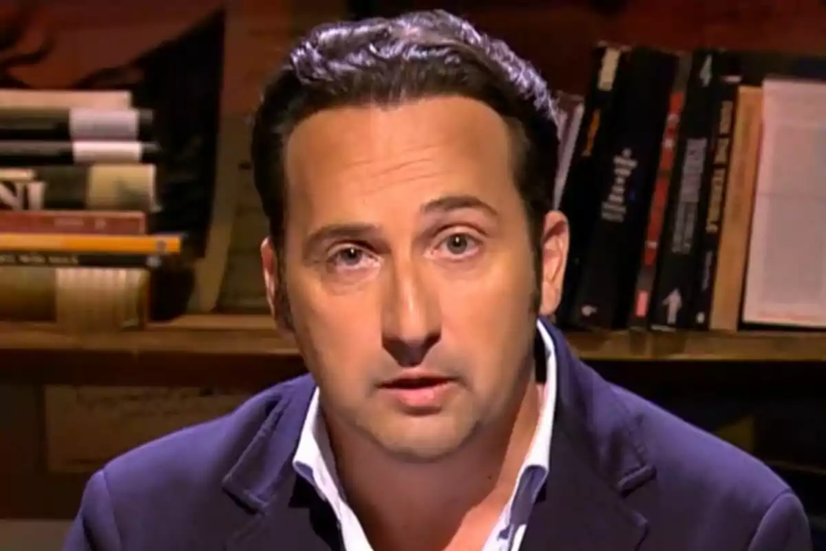 Iker Jiménez de cabello oscuro con chaqueta azul y camisa blanca sentado frente a una estantería con libros en Cuarto Milenio
