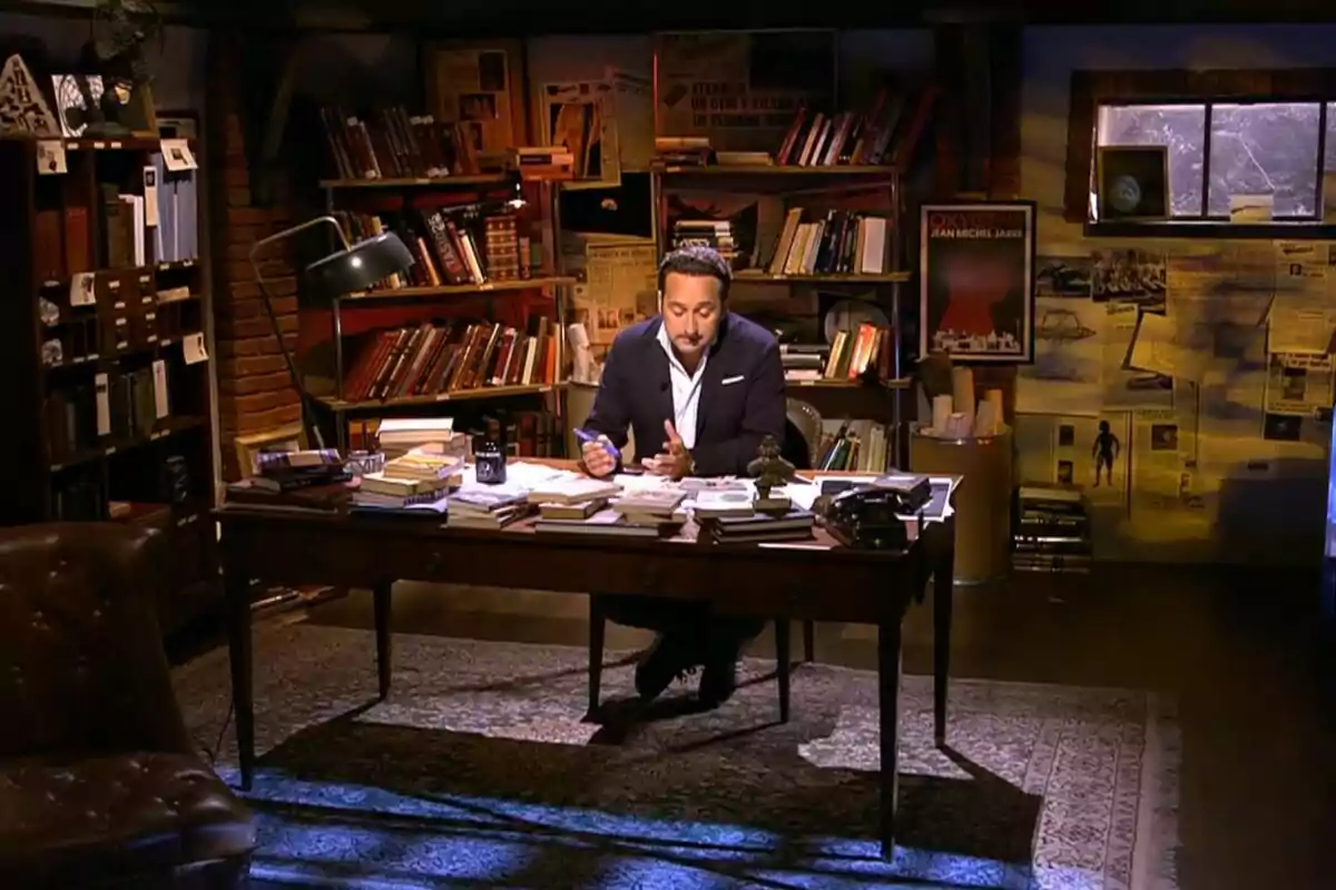 Iker Jiménez sentado en un escritorio antiguo rodeado de libros y papeles en una biblioteca con luz cálida y ambiente acogedor en Cuarto Milenio Iker Jiménez sentado en un escritorio antiguo rodeado de libros y papeles en una biblioteca con luz cálida y ambiente acogedor en Cuarto Milenio