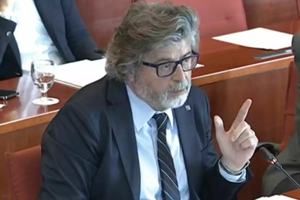 Un hombre con barba y gafas está hablando en una reunión formal mientras gesticula con la mano. Un hombre con barba y gafas está hablando en una reunión formal mientras gesticula con la mano.