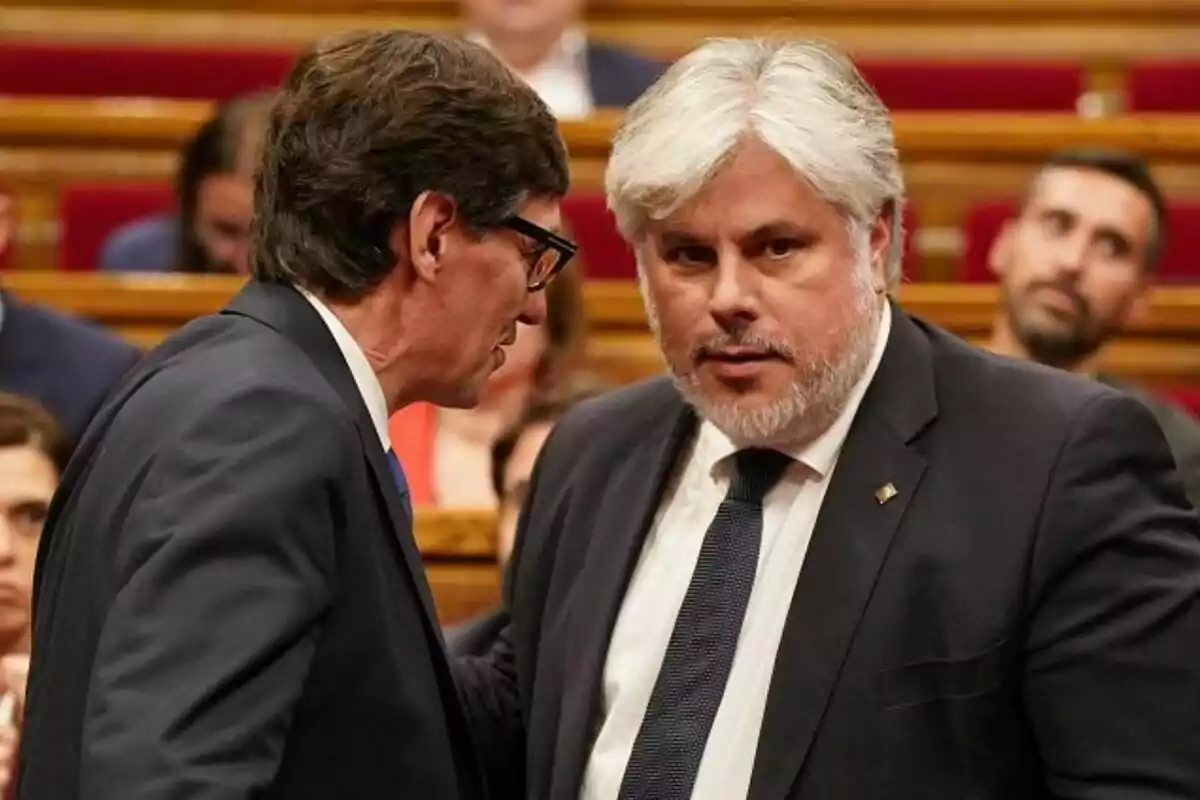 Dos hombres en traje conversan en un entorno formal con personas al fondo.