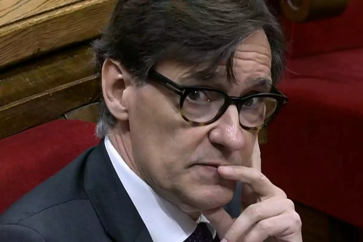 Hombre con gafas de pasta oscuras sentado en un banco rojo, mirando pensativo hacia la cámara y apoyando la mano en su rostro