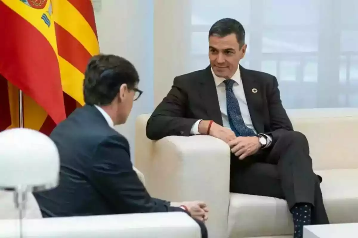 Dos hombres en trajes oscuros conversan sentados en un sofá blanco con banderas de fondo.