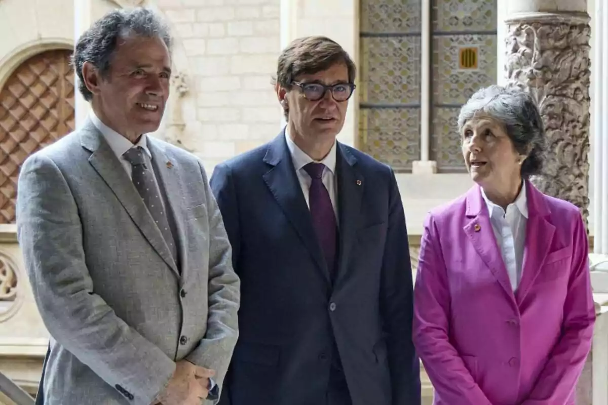 Tres personas de pie en un entorno arquitectónico elegante, con trajes formales y expresiones serias.