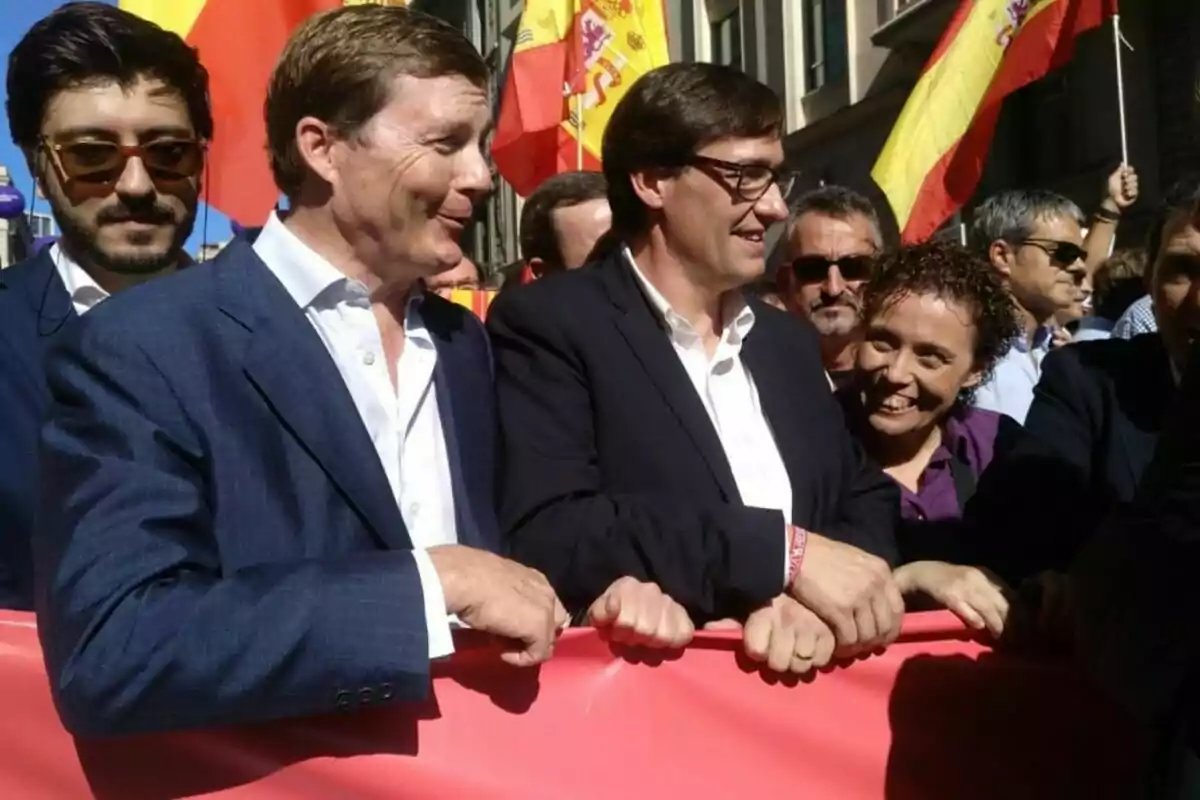Un grupo de personas sonrientes sostiene una pancarta roja en una manifestación con banderas españolas de fondo. Un grupo de personas sonrientes sostiene una pancarta roja en una manifestación con banderas españolas de fondo.