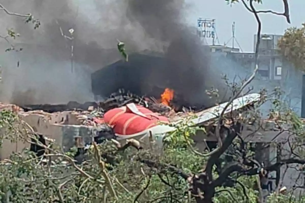 Avión accidentado sobre un edificio con humo negro y fuego saliendo de los restos entre árboles y escombros