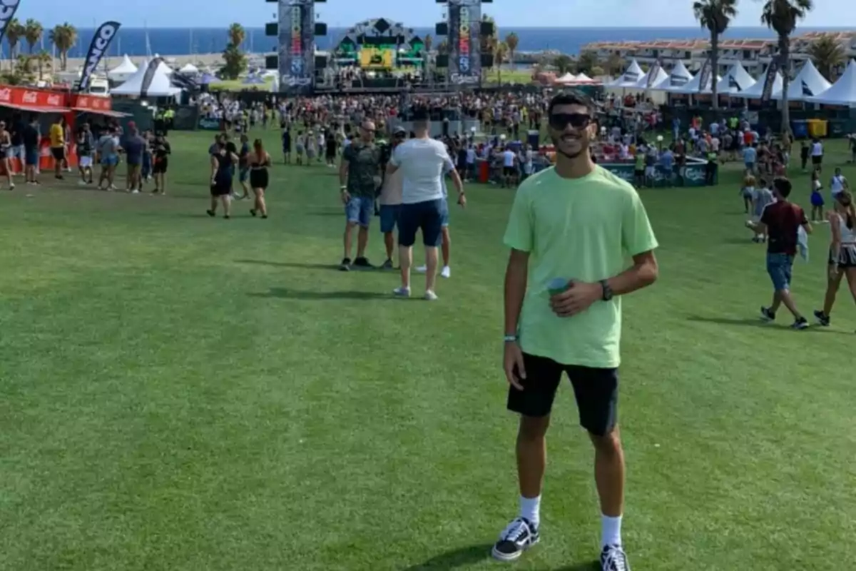 Un hombre con camiseta verde y pantalones cortos negros está de pie en un festival al aire libre con un escenario al fondo y muchas personas alrededor. Un hombre con camiseta verde y pantalones cortos negros está de pie en un festival al aire libre con un escenario al fondo y muchas personas alrededor.