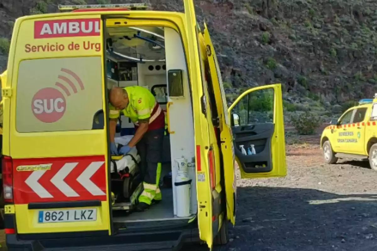 Una ambulancia amarilla con las puertas traseras abiertas muestra a un paramédico atendiendo a una persona en una camilla mientras un vehículo de bomberos está estacionado cerca en un entorno montañoso. Una ambulancia amarilla con las puertas traseras abiertas muestra a un paramédico atendiendo a una persona en una camilla mientras un vehículo de bomberos está estacionado cerca en un entorno montañoso.