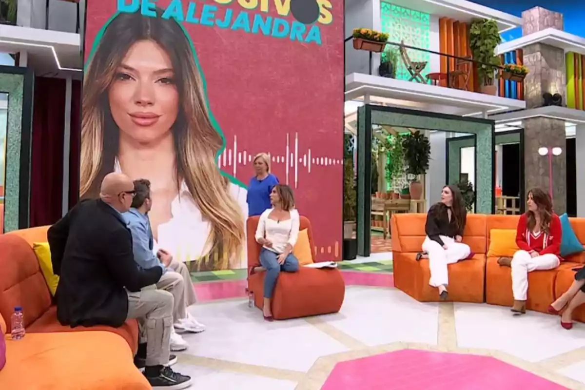 Un grupo de personas está sentado en un set de televisión colorido, con un gran cartel de fondo que muestra la imagen de una mujer y el texto Un grupo de personas está sentado en un set de televisión colorido, con un gran cartel de fondo que muestra la imagen de una mujer y el texto