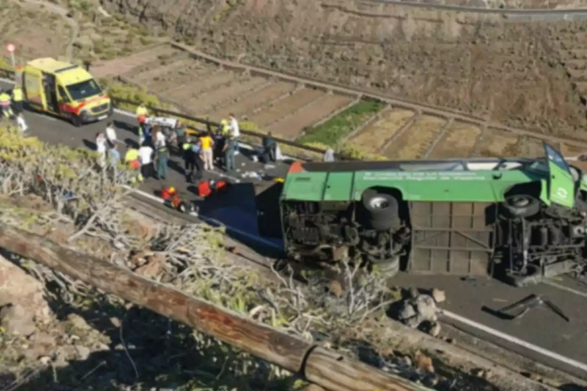 Un autobús volcado en una carretera de montaña con una ambulancia y varias personas alrededor. Un autobús volcado en una carretera de montaña con una ambulancia y varias personas alrededor.