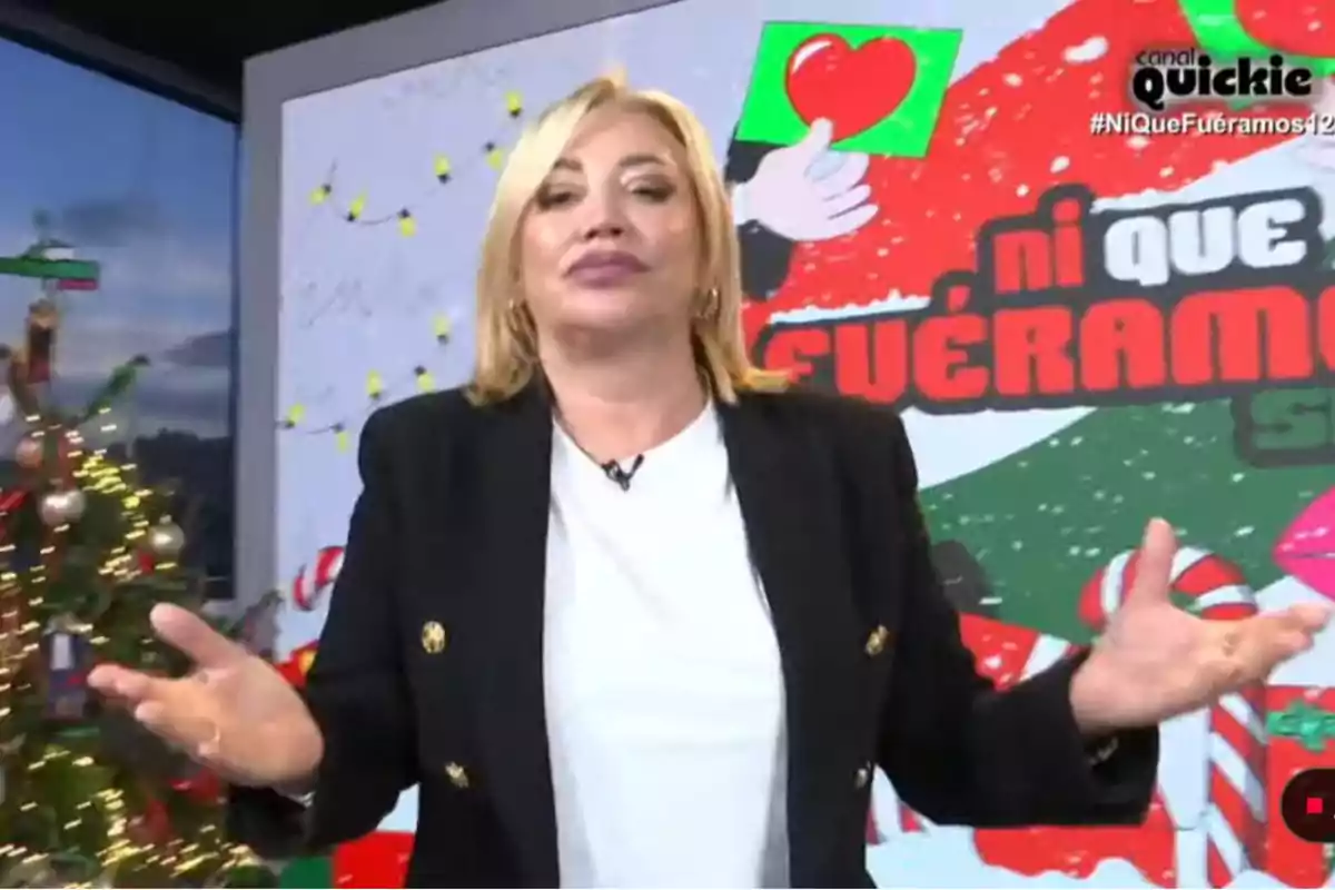 Belén Esteban con cabello rubio y chaqueta negra está frente a un fondo decorado con motivos navideños y un cartel que dice 