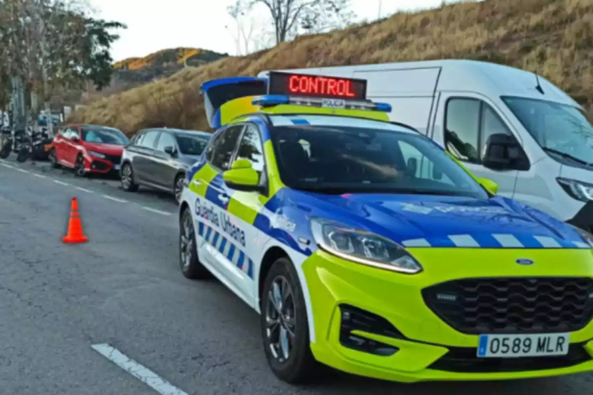 Un coche de la Guardia Urbana de Barcelona con un letrero de control en el techo está estacionado en una carretera junto a otros vehículos y un cono naranja.