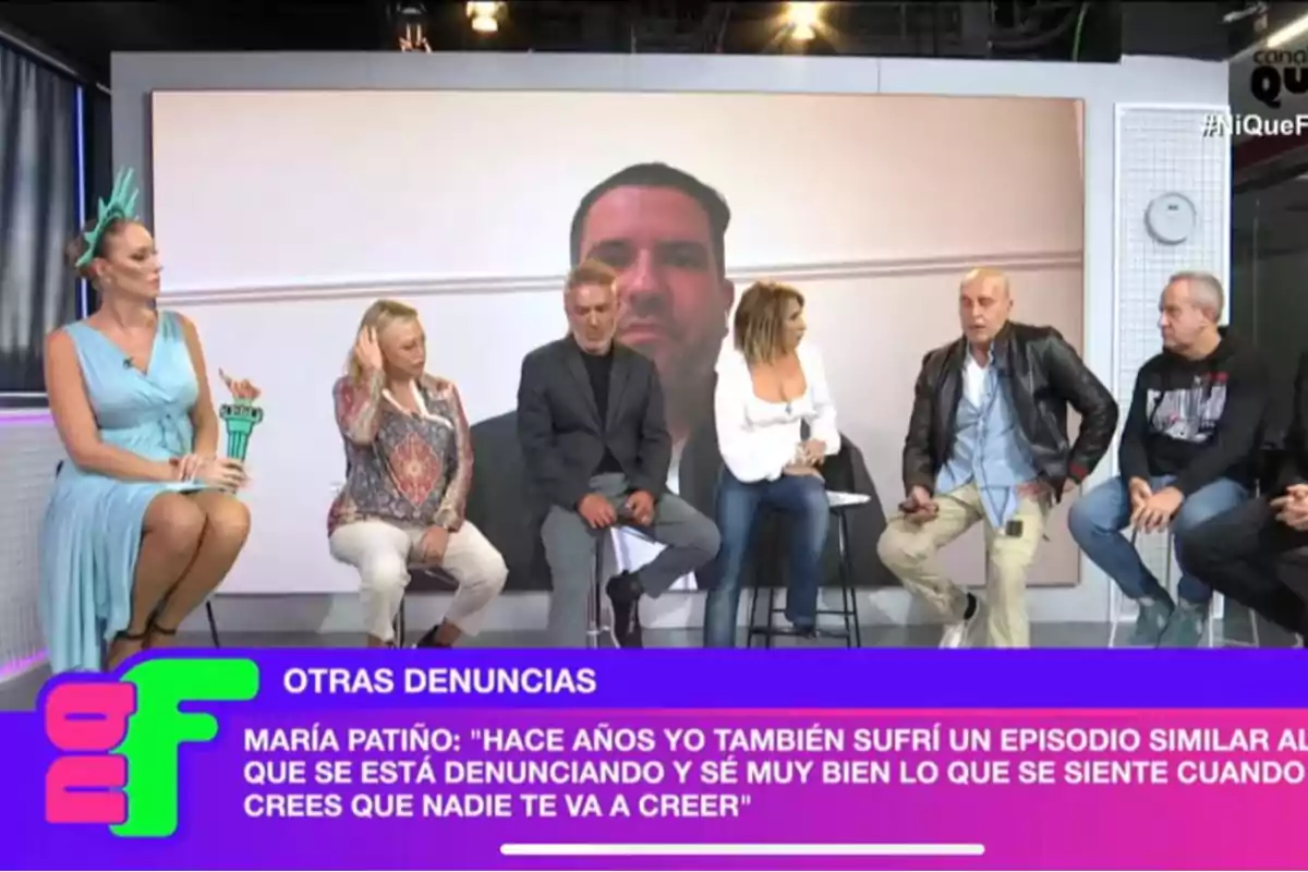 Un grupo de personas sentadas en un set de televisión con un gran rostro proyectado en la pantalla detrás de ellos y un texto en la parte inferior que menciona denuncias y una cita de María Patiño. Un grupo de personas sentadas en un set de televisión con un gran rostro proyectado en la pantalla detrás de ellos y un texto en la parte inferior que menciona denuncias y una cita de María Patiño.
