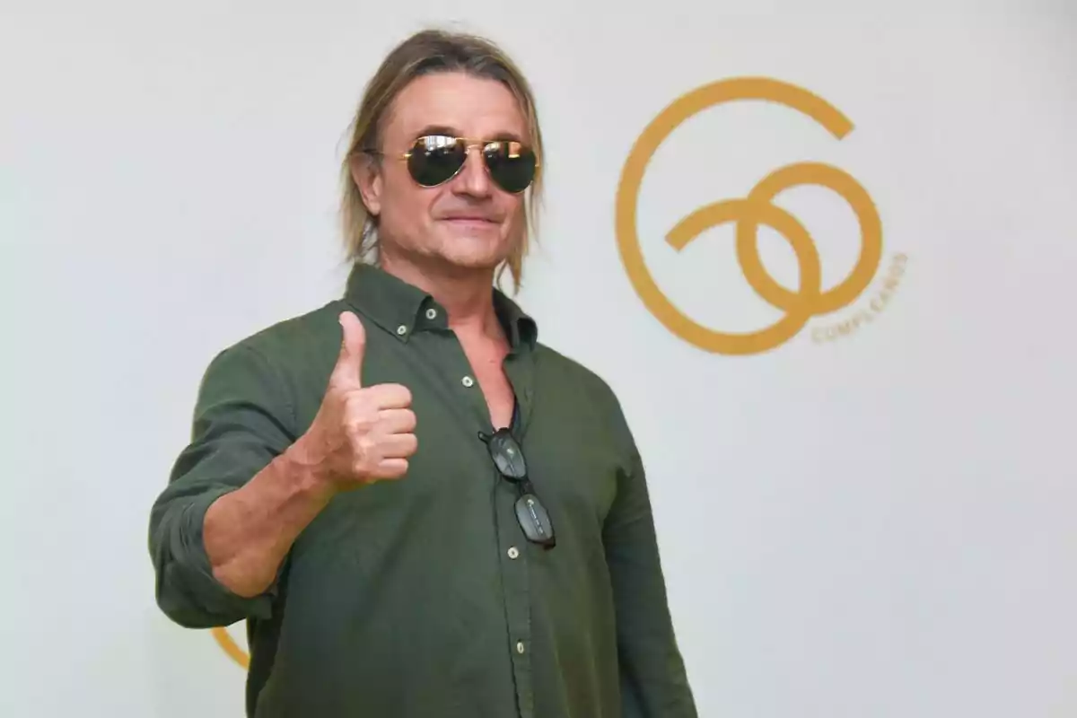 Hombre con gafas de sol y camisa verde haciendo un gesto de aprobación con el pulgar hacia arriba frente a un fondo blanco con un logotipo dorado.