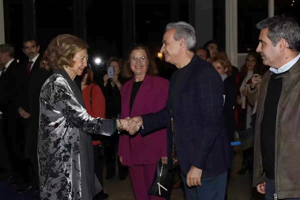 La reina Sofía con un abrigo elegante saluda a un hombre de cabello canoso mientras varias personas observan y toman fotos en el fondo. La reina Sofía con un abrigo elegante saluda a un hombre de cabello canoso mientras varias personas observan y toman fotos en el fondo.