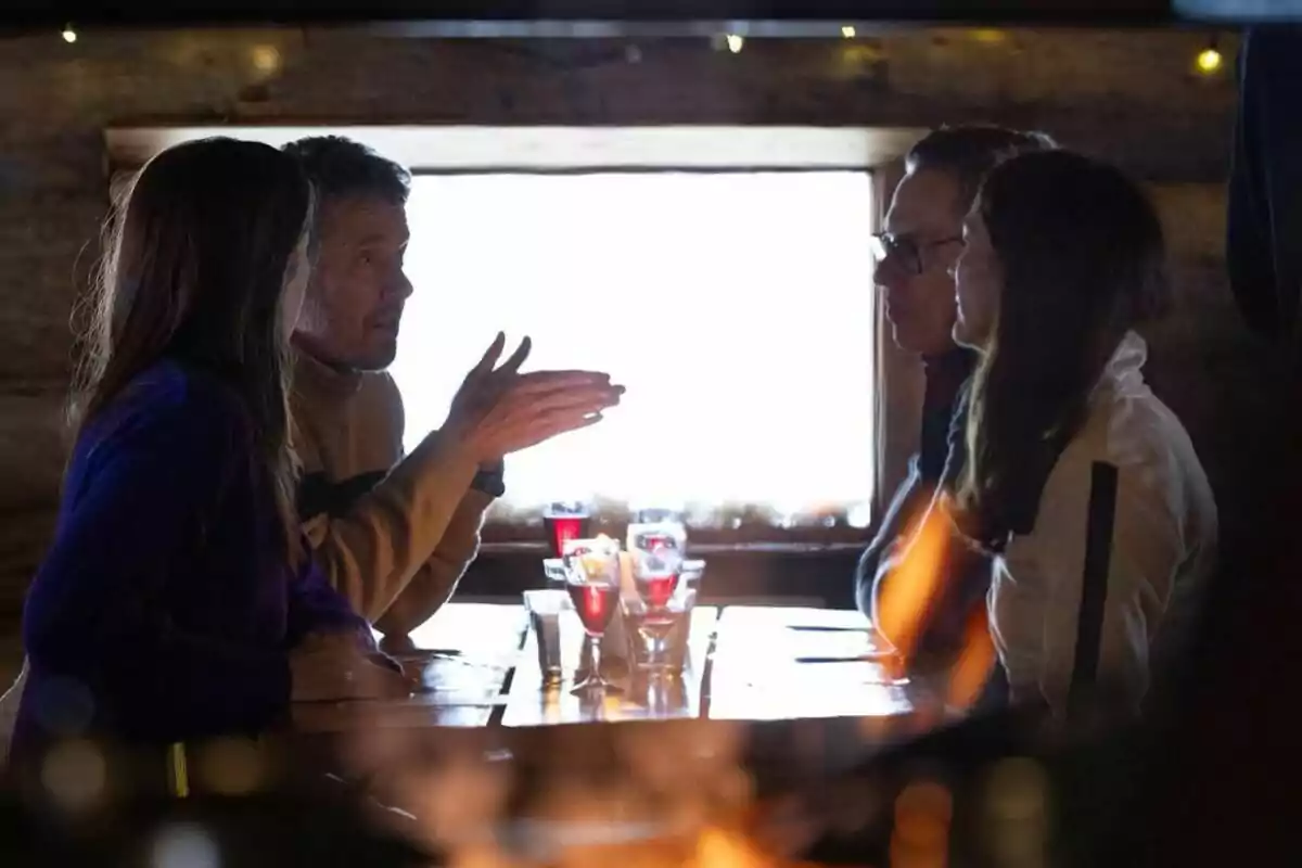 Un grupo de cuatro personas conversando en una mesa de un restaurante con bebidas sobre la mesa y una ventana al fondo. Un grupo de cuatro personas conversando en una mesa de un restaurante con bebidas sobre la mesa y una ventana al fondo.