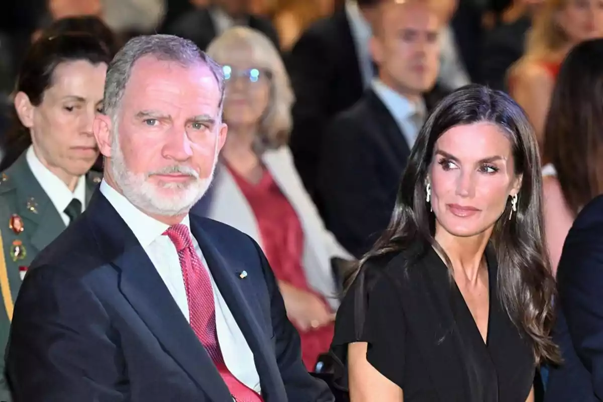 Felipe VI de traje oscuro y corbata roja está sentado junto a la reina Letizia de cabello largo y oscuro vestida de negro en un evento formal rodeados de otras personas Felipe VI de traje oscuro y corbata roja está sentado junto a la reina Letizia de cabello largo y oscuro vestida de negro en un evento formal rodeados de otras personas