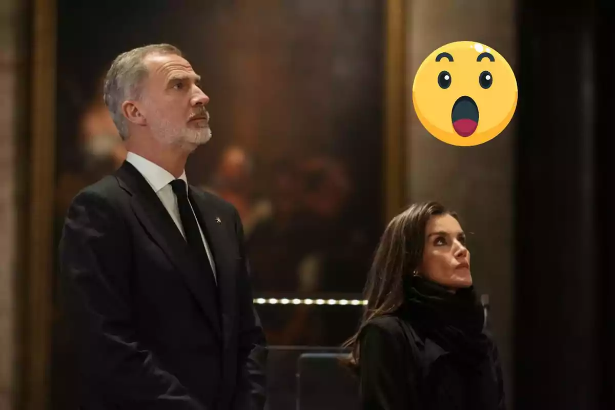 Felipe VI y doña Letizia vestidos de negro mirando hacia arriba con una expresión de sorpresa representada por un emoji.