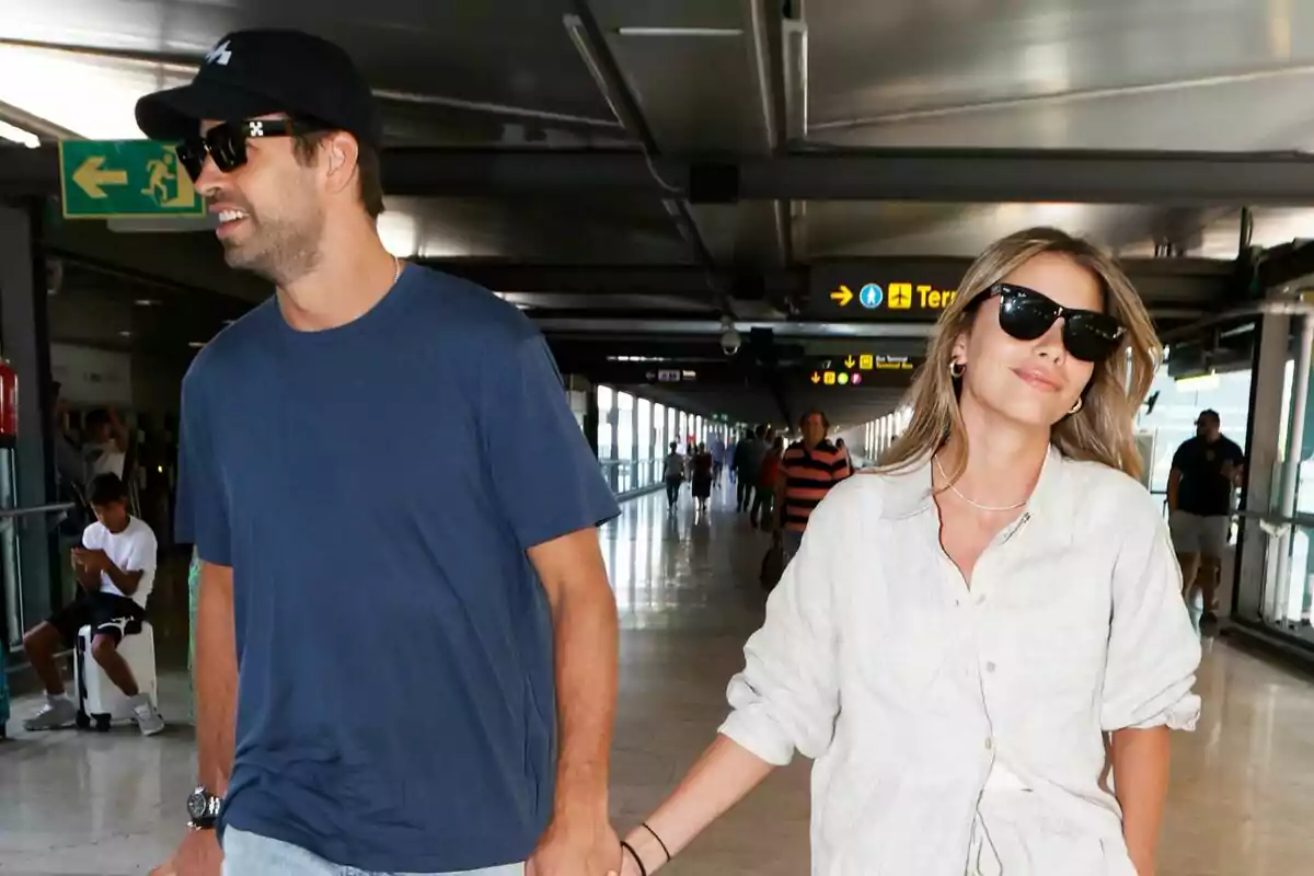 Una pareja camina de la mano en un aeropuerto, ambos llevan gafas de sol y parecen relajados mientras avanzan por el pasillo. Una pareja camina de la mano en un aeropuerto, ambos llevan gafas de sol y parecen relajados mientras avanzan por el pasillo.