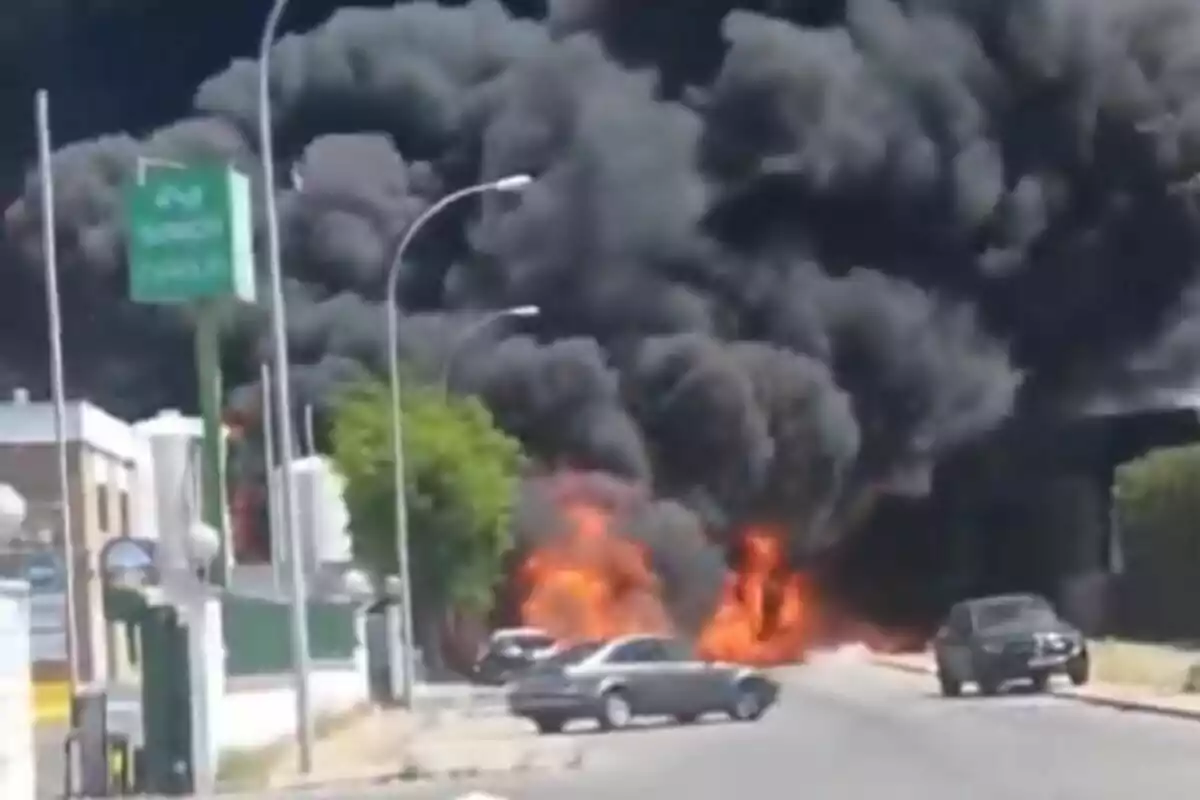 Una gran nube de humo negro se eleva en el cielo mientras las llamas arden intensamente en una calle, con varios coches estacionados cerca y edificios al fondo, en Sevilla.