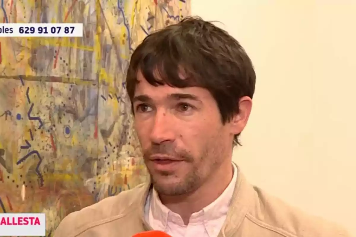 Juan José Ballesta con cabello oscuro y barba ligera está siendo entrevistado frente a una pintura abstracta colorida. Juan José Ballesta con cabello oscuro y barba ligera está siendo entrevistado frente a una pintura abstracta colorida.