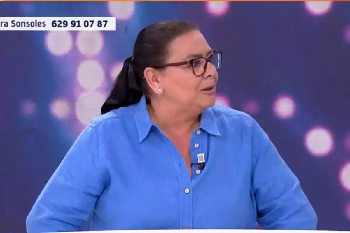 María del Monte con camisa azul y gafas está participando en un programa de televisión con un fondo de luces desenfocadas. María del Monte con camisa azul y gafas está participando en un programa de televisión con un fondo de luces desenfocadas.