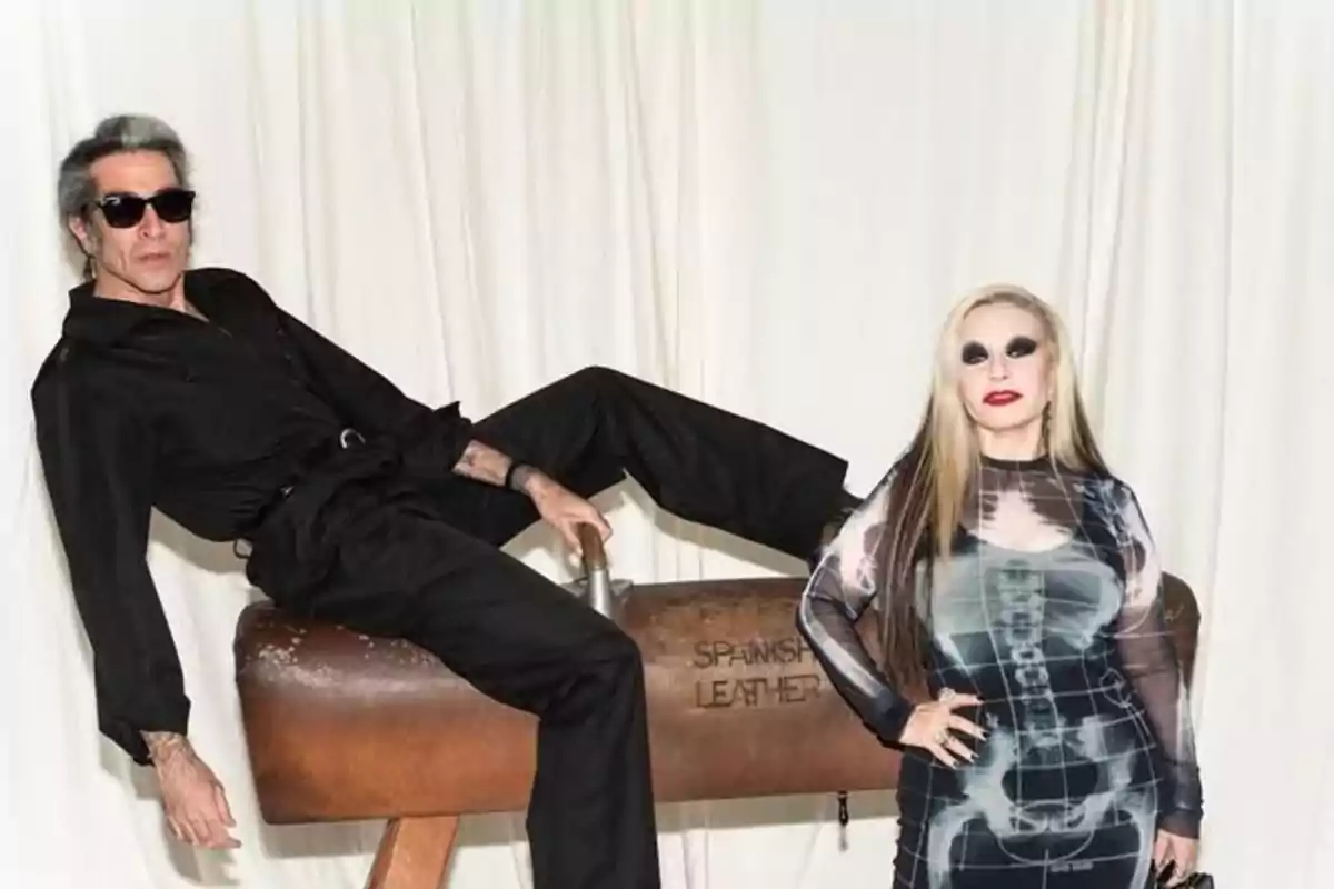 Mario Vaquerizo vestido de negro está recostada sobre un potro de gimnasia mientras Alaska con un vestido estampado está de pie a su lado, ambos posan frente a una cortina blanca. Mario Vaquerizo vestido de negro está recostada sobre un potro de gimnasia mientras Alaska con un vestido estampado está de pie a su lado, ambos posan frente a una cortina blanca.