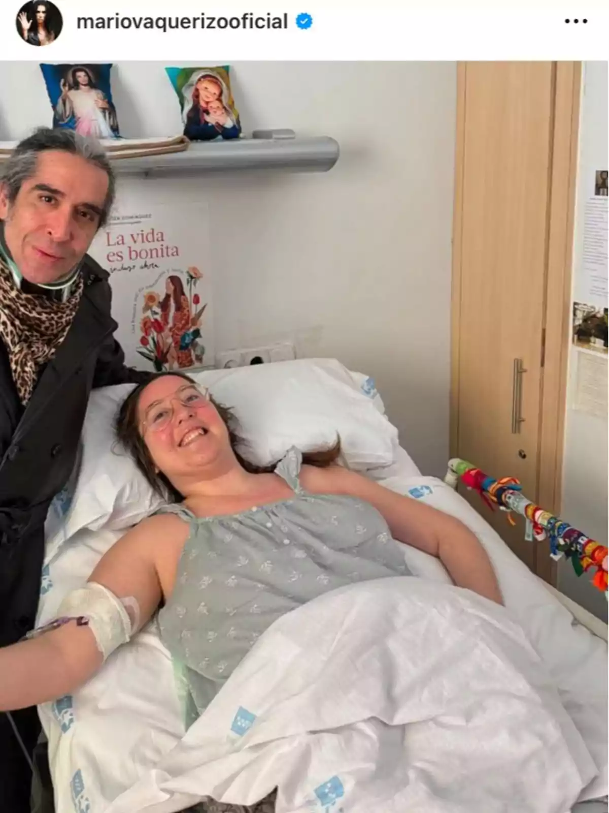 Una mujer sonriente está acostada en una cama de hospital mientras Mario Vaquerizo está de pie a su lado, ambos parecen estar contentos. Una mujer sonriente está acostada en una cama de hospital mientras Mario Vaquerizo está de pie a su lado, ambos parecen estar contentos.