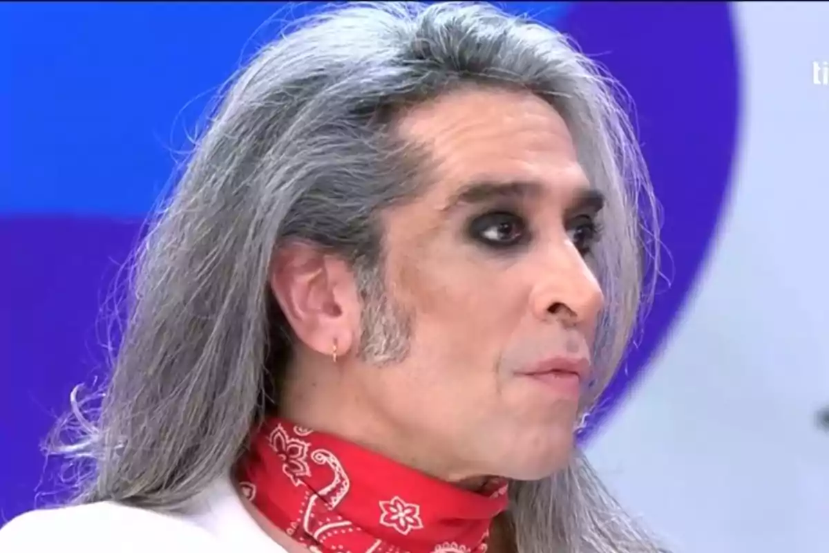 Mario Vaquerizo con cabello largo y canoso, maquillaje oscuro en los ojos y una bandana roja alrededor del cuello. Mario Vaquerizo con cabello largo y canoso, maquillaje oscuro en los ojos y una bandana roja alrededor del cuello.
